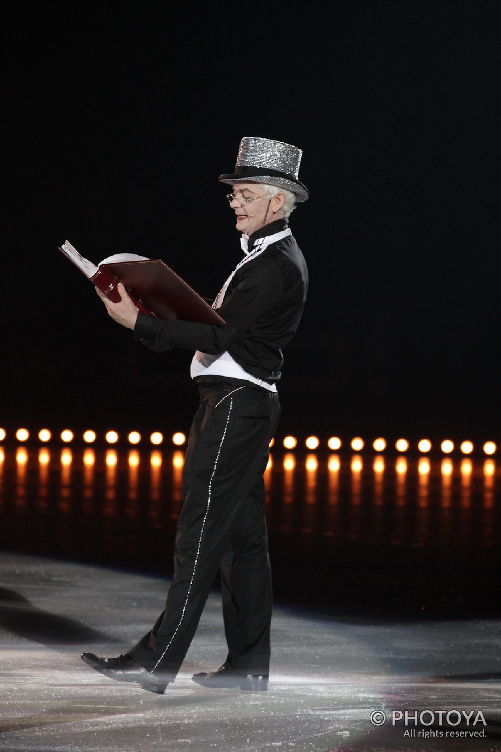 Man dressed in a top hat and tux reading a large tome on stage